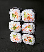 Tasty sushi rolls with rice, avocado and fish are neatly stacked on a dark stone surface, highlighting the intricate textures and bright colours of each ingredient.