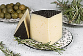 A piece of hard cheese is skilfully presented on a patterned ceramic plate, accompanied by fresh rosemary and green olives. The textured cheese rind adds an elegant touch.