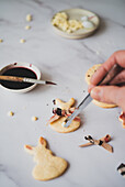 Seen from above, an unrecognisable person uses a thin brush to paint biscuits in the shape of an Easter bunny with hibiscus tea, which lie next to a bowl of bright dark red tea and scraps of dough.