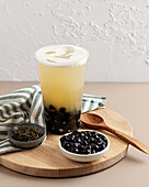 A refreshing cup of bubble tea with creamy foam and tapioca pearls on a wooden board, accompanied by a bowl of pearls, loose tea and a wooden spoon