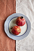 Top view of two ripe pomegranates elegantly placed on a decorative white plate with blue stripes against a textured background of contrasting fabrics.