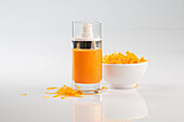 A bright orange-coloured natural oil for skin care in a glass bottle next to a bowl of marigold flowers on a simple white surface with an elegant design.