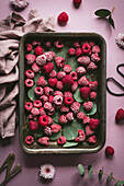 A tray of frozen raspberries with scattered eucalyptus leaves on a pink-coloured surface, surrounded by flowers and fabric, creates a natural and rustic setting.