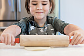 A smiling child wearing an apron with a star pattern rolls out dough with a wooden rolling pin in a brightly lit kitchen. Flour on the nose adds a playful touch to the baking activity.