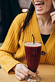 A woman in a cosy yellow jumper holds a tall glass of coffee with a black straw in her hand and smiles joyfully The image captures a moment of refreshment and happiness