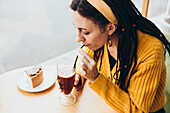 A woman with dreadlocks in a café sips a drink through a straw. Wearing a yellow jumper and a headband, she enjoys a slice of cake on a light wooden table that creates a cosy, warm atmosphere