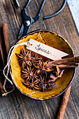 Top view of star anise and cinnamon sticks in a decorative yellow bowl with a sign labelled Spices, surrounded by rustic tools on a wooden surface.