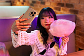 An Asian Gen Z girl takes a selfie with her mobile phone while enjoying candyfloss in a lively arcade, combining fun and modern youth culture