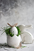A carefully crafted Easter decoration with a white speckled egg lying in a nest of fresh green grass, topped with a delicate feather, and an elegant white ceramic bunny nearby, against a soft, textured grey background.