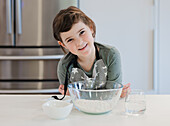 A small child in an apron smiles while preparing to bake in a modern kitchen. The scene shows a glass bowl of flour and a glass of water nearby.