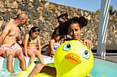 A cheerful girl enjoys a sunny day at the swimming pool and sits on a yellow inflatable duck while her family relaxes nearby. Perfect summer fun, family time and outdoor activity.