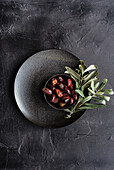 Top view of a dark textured plate filled with shiny Greek grilled olives, garnished with fresh green olive leaves that have a rustic and appetising appearance, ideal for Mediterranean cuisine themes.