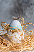 An Easter egg in soft blue and with natural feathers, nestled in a rustic straw nest, is a traditional Easter nest with a touch of elegance.