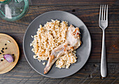 A tasty dish of cooked rice with a tender leg of rabbit, served on a grey plate The rustic wooden placemat includes a fork, garlic and pepper for added flavour