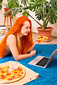 A person with lively hair enjoys a slice of pizza while surfing on a laptop on a blue plush carpet Houseplants and books create a cosy atmosphere in the house