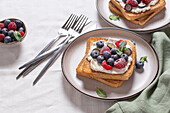 A delicious portion of toast with cream cheese and fresh raspberries and blueberries, beautifully arranged on a chic white tablecloth, next to silver cutlery and a small bowl with additional berries.