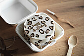 South Korean bento birthday cake with 'Happy Birthday' lettering and chocolate decorations, presented in a white takeaway box, along with a wooden spoon and a matte ribbon.