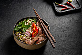 Traditional Vietnamese beef pho noodle soup, served in a stylish dark bowl, garnished with slices of beef, red chilli, onions and fresh herbs on a dark background.