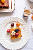 Top view of a gourmet dessert scene on a light-coloured tablecloth with a piece of cake topped with cream and fresh berries, cherries, a small apricot and a shot of espresso.