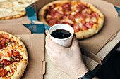 Cropped, unrecognisable male hand holding a soda drink in a cup against a blurred background of cardboard boxed pizzas suggesting a casual dining environment.