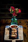 Crispy spring rolls lie on a wooden board next to a bowl of dipping sauce, framed by a bright bouquet of red flowers in a transparent vase, creating a cosy atmosphere