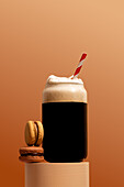 A refreshing coffee in a glass with foam and a red striped straw, accompanied by two delicious macarons The warm, brown background adds a cosy touch