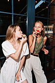 Two friends share a fun moment while eating slices of pizza baked in a wood-fired oven. Casual al fresco dining with fresh, tasty pizza and good humour in a modern setting.