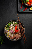 Top view of a delicious bowl of Vietnamese beef pho noodle soup with tender slices of beef, flat rice noodles, fresh herbs such as coriander and basil and vibrant garnishes such as lime, red chilli and sprouts against a textured dark background