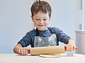 A small child in an apron with a star pattern happily rolls out dough with a wooden rolling pin. His nose is dusted with flour, expressing the joy of baking in the kitchen at home.