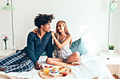 A young couple sits happily on a bed and shares a breakfast tray with fruit, juice and pastries. The room is well lit and creates a warm, intimate atmosphere.
