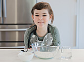 A cheerful child in an apron smiles while preparing to bake in a modern kitchen. The picture shows clear bowls of flour and water, depicting a delightful cooking scene.