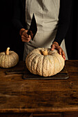 A person in an apron prepares to cut a large pumpkin on a wooden table. The warm, rustic setting highlights the natural texture of the pumpkins and creates an autumnal atmosphere.