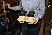 A person opens the door of a café holding a tray with four takeaway coffee cups. The scene conveys a warm, inviting atmosphere and emphasises the theme of coffee specialities.