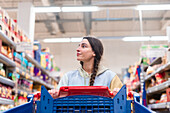 A woman with a braided ponytail looks up as she pushes a red shopping trolley in a supermarket aisle.