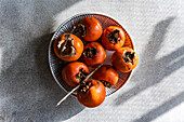 A close-up view from above of freshly picked ripe persimmons neatly arranged on a stylish white and blue striped plate. The fruits have a rich orange-reddish colour under soft, natural light that highlights their freshness.