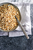 Top view of a bowl filled with finely chopped hazelnuts and a spoon against a textured grey background that highlights texture and natural colours.