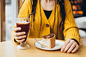 Close-up of a person in a yellow jumper holding a glass with a drink and a plate with a piece of cake in a cosy café - perfect for depicting relaxation