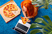 A person with colourful hair enjoys a slice of pizza while using a laptop on a bright blue carpet Green leaves frame the cosy indoor scene, which is perfect for leisure or remote working