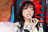 An Asian Gen Z girl enjoys candyfloss in a bright, colourful arcade She shows a sense of playful joy, surrounded by colourful lights and excitement