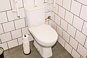 Eine saubere weiße Toilette in einem Café, umgeben von weißen U-Bahn-Fliesen und einem grauen Boden. In der Nähe befindet sich ein Stapel Toilettenpapierrollen, um die Hygiene und den Komfort der Kunden zu gewährleisten.