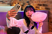 Asian Gen Z girl takes a selfie while enjoying candyfloss in an arcade The bright lights and playful atmosphere capture youthful energy and modern culture