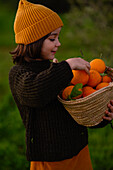 A small child in a hat and dark jumper smilingly holds a basket of fresh oranges in a lush, green outdoor setting, capturing a moment of joy and connection with nature.