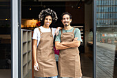Two café employees in matching brown aprons stand side by side at the entrance to their café, demonstrating teamwork and customer service in a welcoming environment