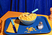 A bowl of creamy macaroni and cheese stands on a blue tray with a fork. A salt shaker, a knife and a dotted napkin complete the colourful, inviting table decoration.