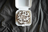 Top view of a bento birthday cake with Happy Birthday lettering in a white takeaway box against a grey, textured background.