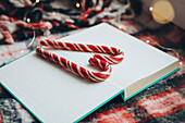 Red and white striped candy canes stylishly arranged on an open book, surrounded by soft, festive woollen fabric and twinkling fairy lights, expressing the joy and warmth of the Christmas season.