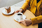 A person in a yellow jumper sits at a wooden table enjoying a cup of coffee next to a piece of cake The scene evokes warmth and relaxation