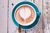 Close-up of a heart-shaped latte motif, served in a turquoise-coloured cup on a worn wooden table. Perfect for concepts of warmth and comfort.