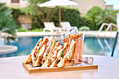 A delicious sandwich with ham, cheese and tomatoes, served with crispy chips in a basket. The setting is peaceful, by a pool, and evokes relaxation and pleasure.