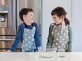 Two siblings in aprons with a star pattern smile at each other in a kitchen. A mixing bowl with flour and a cup of water stand on the counter, creating a warm, playful atmosphere.